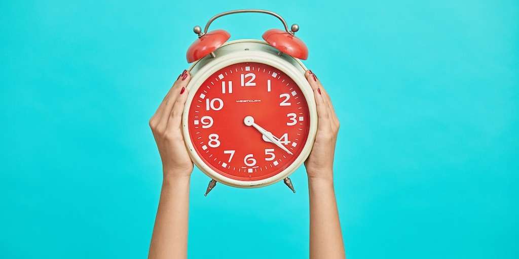 Hands holding a large red alarm clock against a bright turquoise background, symbolizing the concept of 'Time Inversion'.