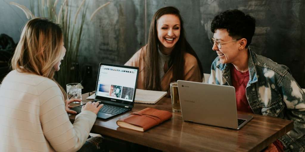 Three colleagues laughing, sharing a mindset change discussion over laptops at a rustic coffee shop table.