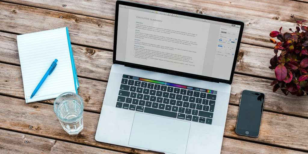 Open laptop displaying document on wooden desk, keyphrase Best Ways to Be Productivity, with notepad, pen, and smartphone.