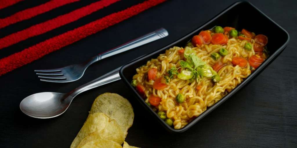 A bowl of 2 Min Noodles with peas, carrots, and greens, served with a side of chips.