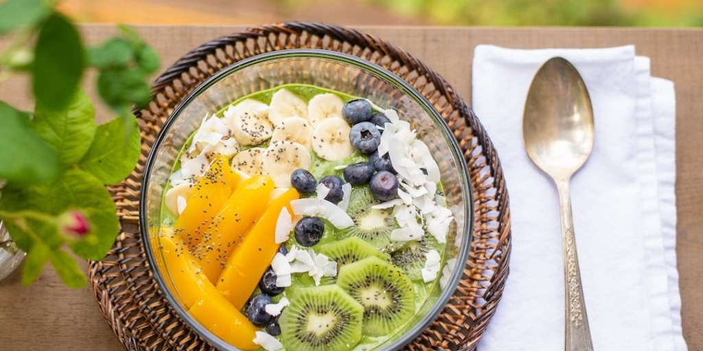 A vibrant bowl of Spirulina Crunchies Superfood breakfast bowl, topped with sliced mango, kiwi, banana, blueberries, coconut flakes, and chia seeds, ready to be enjoyed with a silver spoon on the side.