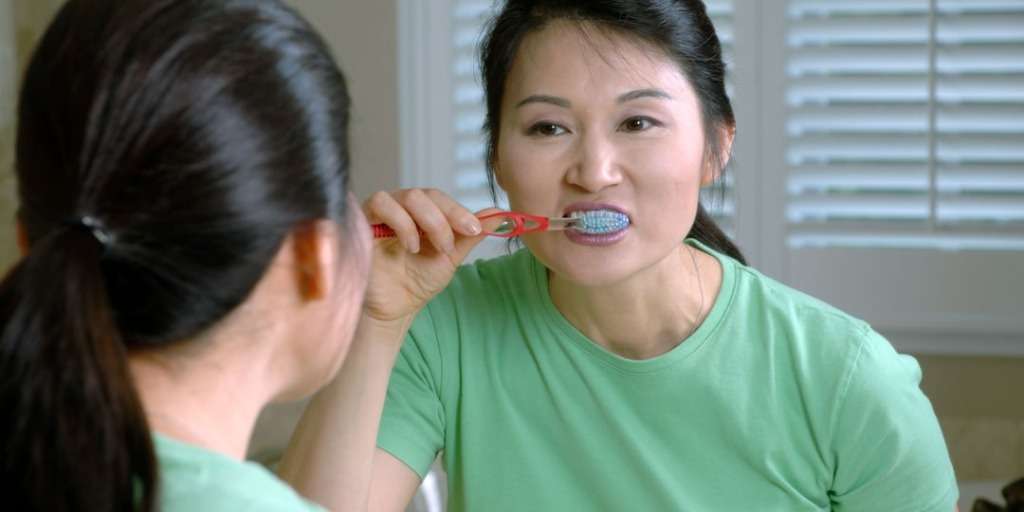 Woman in green shirt brushing teeth, reflecting on timing your brushing after meals in mirror.