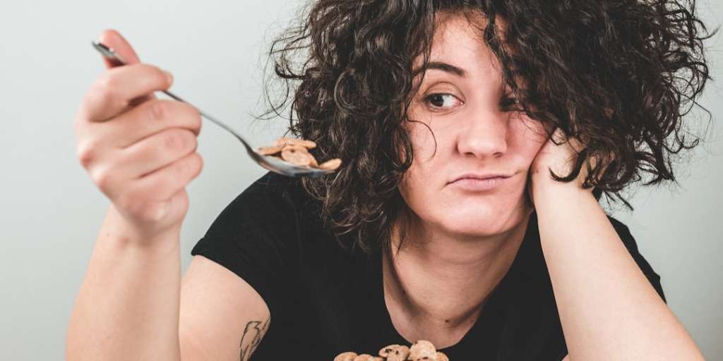 Woman with curly hair looking bored while eating cereal, considering ways to reduce cortisol.