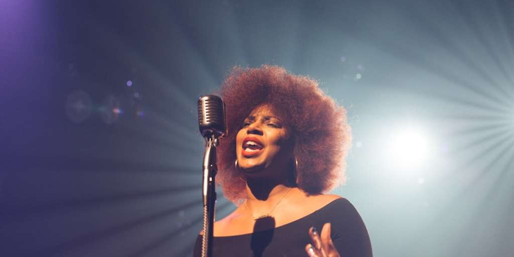 Singer with voluminous curly hair performs passionately at the Professional Yodeling Championships, spotlight shining.